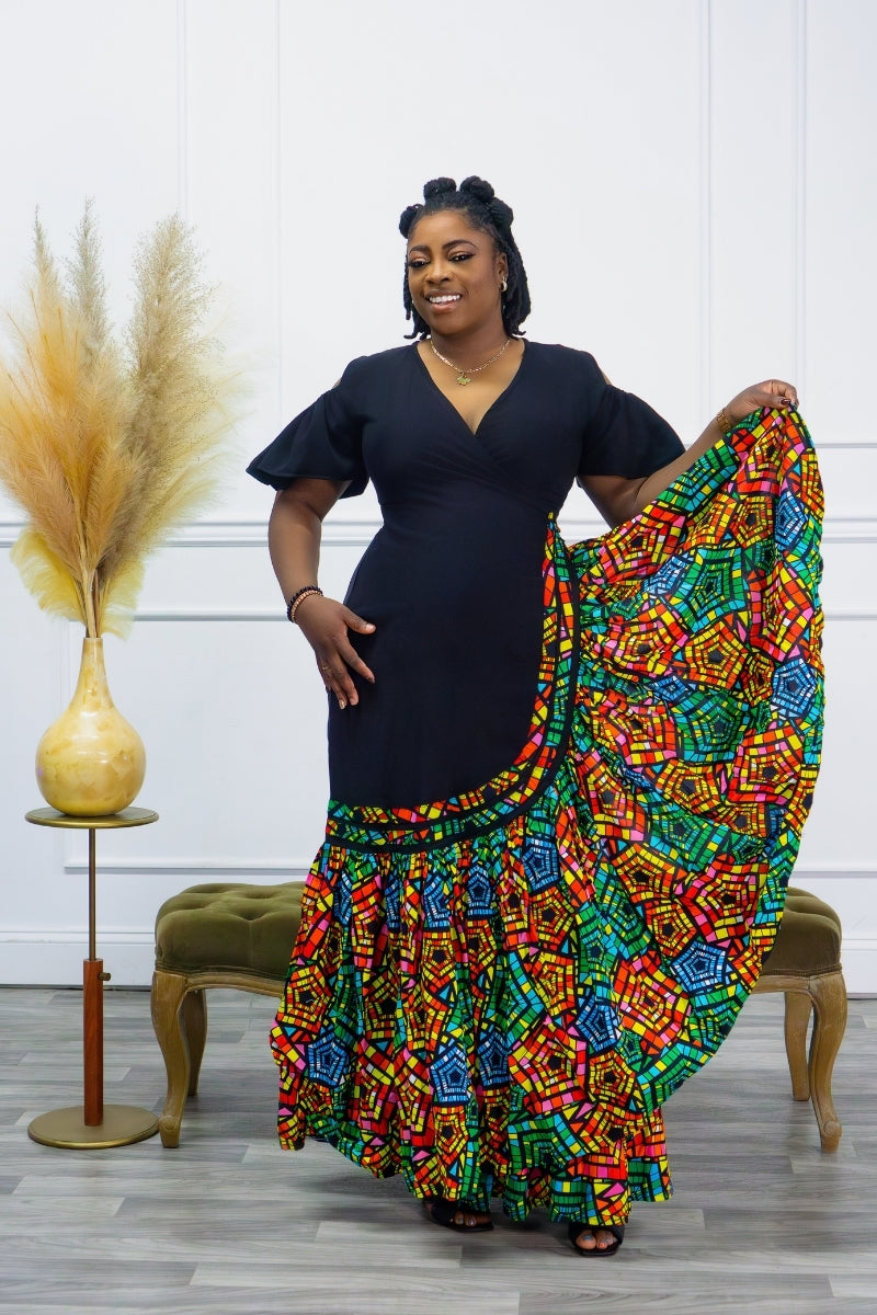 A woman standing and posing in a colorful, geometric-patterned maxi gown with cut-out sleeves. The gown has a fitted waist and flows out at the bottom, with a vibrant mix of colors including red, blue, and green.