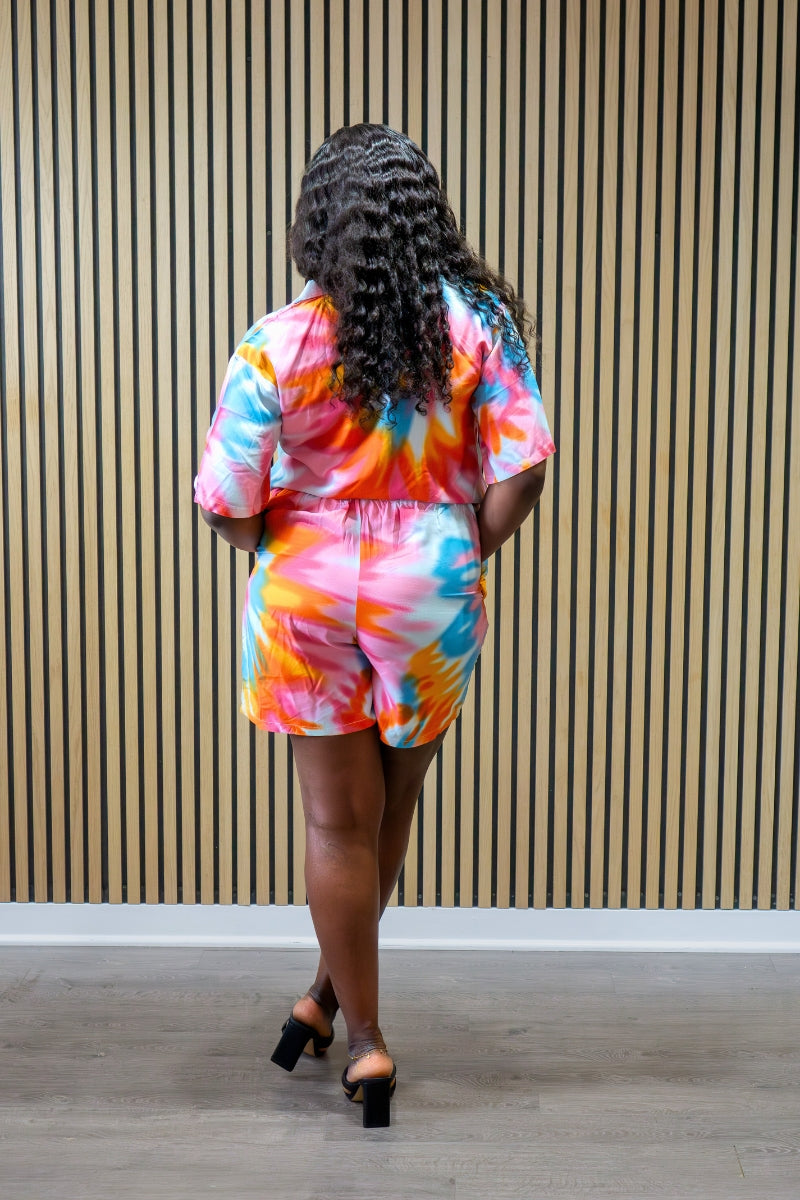 Person wearing a colorful tie-dye outfit standing against a striped wall.