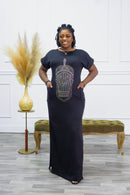 A woman standing in a room with a black, sleeveless, maxi dress with a geometric pattern on the chest area, featuring pockets on the sides and a slit. She has rolled short sleeves and the dress is on a crepe fabric.