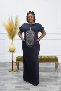 A woman standing in a room with a black, sleeveless, maxi dress with a geometric pattern on the chest area, featuring pockets on the sides and a slit. She has rolled short sleeves and the dress is on a crepe fabric.
