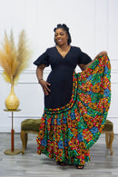 A woman standing and posing in a colorful, geometric-patterned maxi gown with cut-out sleeves. The gown has a fitted waist and flows out at the bottom, with a vibrant mix of colors including red, blue, and green.