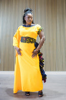 A woman modeling a vibrant mustard-yellow maxi dress with asymmetrical sleeves, a decorative Ankara-print waistband, and side ruffle detailing.