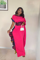 A woman modeling a vibrant mustard-yellow maxi dress with asymmetrical sleeves, a decorative Ankara-print waistband, and side ruffle detailing.