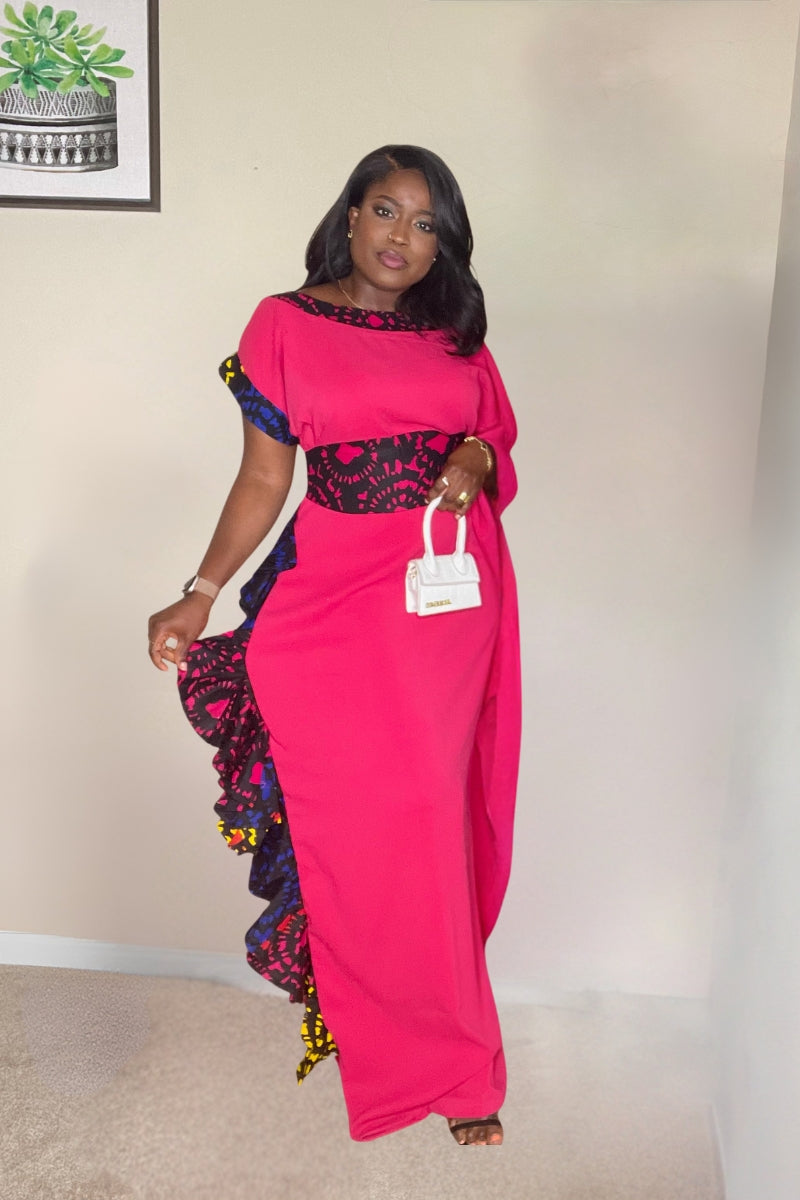 A woman modeling a vibrant mustard-yellow maxi dress with asymmetrical sleeves, a decorative Ankara-print waistband, and side ruffle detailing.