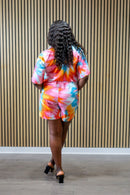 Person wearing a colorful tie-dye outfit standing against a striped wall.